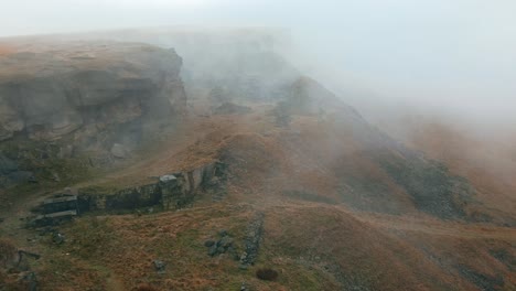 Aerial-view-of-clouds-moving-slowly-over-the-hills,-Pennines-on-a-foggy-morning,-golden-hills-and-beautiful-rocky-cliffs-and-moorlands