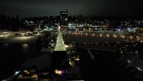 Winter-night-drone-shot-of-a-small-town-with-Christmas-lights-and-snow-over-lake