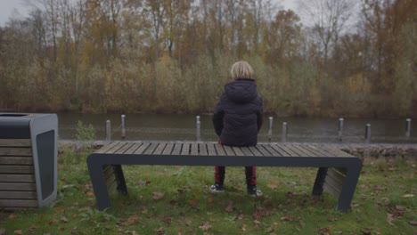 Teenage-boy-sitting-down-on-park-bench-overlooking-river-in-autumn
