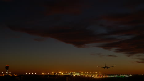 Avión-Aterrizando-Al-Atardecer,-Aeropuerto-De-Barcelona-Iluminado-En-Segundo-Plano
