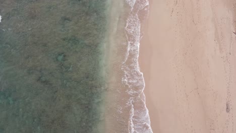 Stable-Aerial-top-view-over-ocean-water-at-the-shore,-washing-the-golden-sand-of-the-beach-with-footprints,-summer-vacation-on-Hawaii-island