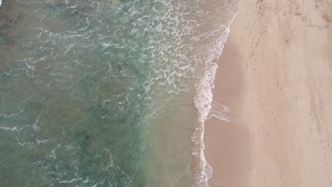 Aerial-top-view-over-clear,-Turquoise-ocean-water-foaming-at-the-shore,-washing-the-golden-sand-of-the-beach-with-footprints,-summer-vacation-on-Hawaii-island