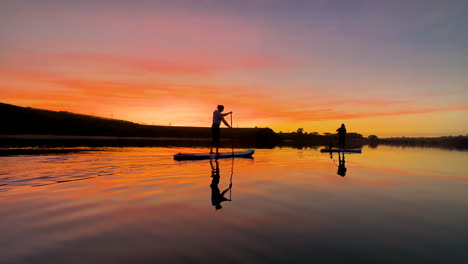 Two-silhouettes-of-a-couple-girsl-enjoying-a-beautiful-sunset-paddleboard-with-an-awesome-water-refelction-in-southern-california-on-a-local-lagoon-in-carlsbad,-ca