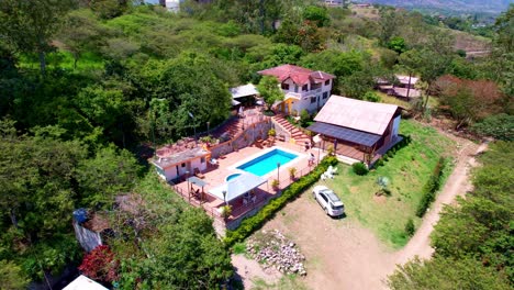 Aerial-shot-with-drone-from-the-view-of-a-hacienda-with-a-red-roof-and-white-walls-surrounded-by-large-leafy-and-green-trees-with-a-blue-pool-and-a-sunny-summer-day