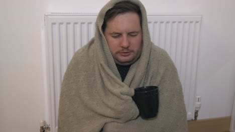 Freezing-young-man-wrapped-in-a-blanket-sitting-next-to-a-heater,-shivering-from-cold-and-drinking-tea