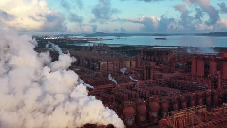 Drone-shot-of-coal-and-metal-factory-at-sunrise,-flying-near-large-steam-smoke-stacks-in-operation