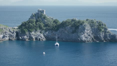 Catamaran-Sailboat-And-Small-Boats-Docked-Near-Rocky-Headland-With-Torre-del-Capo-di-Conca-Atop-On-The-Amalfi-Coast-In-Italy