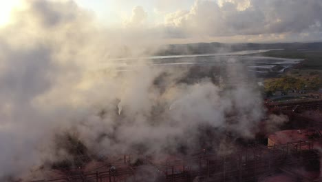 Drone-shot-of-coal-and-metal-factory-at-sunrise,-flying-through-large-steam-smoke-stacks-in-operation