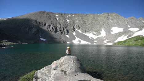 Lonely-Woman-Sitting-on-Rock-in-Front-of-Idyllic-Alpine-Lake-Under-Mountain-Hills-on-Sunny-Summer-Day