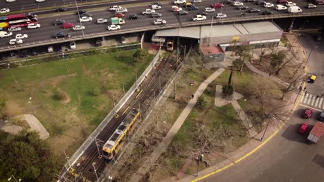 Aerial---Top-view-of-yellow-tram-arriving-station-and-cars-on-road-overhead-in-Buenos-Aires