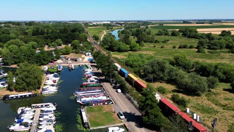 Aerial-view-of-a-long-goods-train-passing-by-a-marina-at-Ely,-Cambridgeshire,-UK