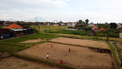 Los-Niños-Juegan-Al-Fútbol-En-El-Campo-De-Pasto-Timelase-Antena-Al-Aire-Libre-Pueblo-Rural,-Bali,-Indonesia,-Parque-Vacante-En-Campos-De-Arroz