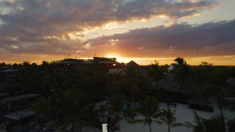 Aerial-view-of-the-sun-setting-behind-the-tulum-landscape-in-Mexico-while-tourists-walk-over-the-Akiin-Beach