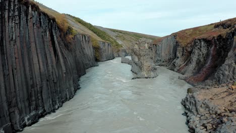 Aerial-down-glacial-river-with-dramatic-basalt-rock-columns,-Studlagil,-Iceland