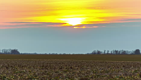Static-shot-of-brown-agricultural-field-with-sun-setting-in-the-background-on-a-cloudy-day-in-timelapse