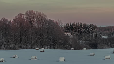 Shot-of-hay-bales-covered-with-snow-over-agricultural-farmlands-winter-evening-with-fog-passing-by-in-timelapse