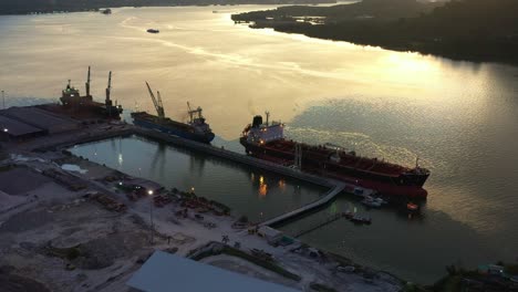 Birds-eye-view-fly-around-Lumut-port-industrial-park-along-sungai-manjung-river-with-commercial-bulk-carrier-ships-docked-at-the-port,-tilt-up-reveals-mountain-landscape-at-sunset,-Perak,-Malaysia