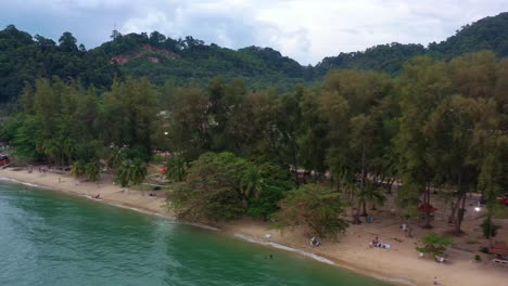 Cinematic-low-level-aerial-drone-fly-along-coastal-shore-of-touristic-destination-teluk-batik-beach-surrounded-by-lush-green-trees-in-a-tropical-atmosphere-at-Lumut,-Perak,-Malaysia,-Southeast-Asia
