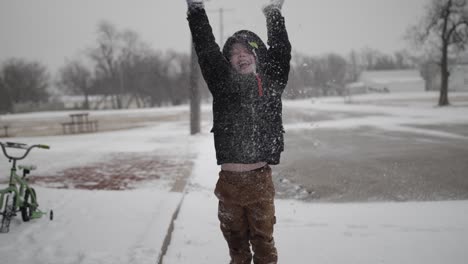 Young,-happy,-boy-smiling-and-wearing-a-coat-playing-outside-on-a-cold,-winter-day-in-December-throws-snow-in-the-air-during-Christmas-break-in-a-small-town-in-the-midwest