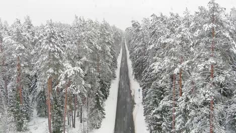 Magical-Christmas-road-and-woodland-covered-in-snow-during-snowfall,-aerial-view