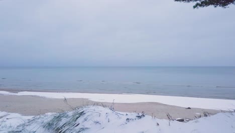Beautiful-aerial-establishing-view-of-Baltic-sea-coast-on-a-overcast-winter-day,-beach-with-white-sand-covered-by-snow,-coastal-erosion,-climate-changes,-wide-angle-drone-shot-moving-backward