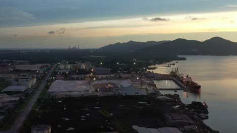 Aerial-tracking-shot-capturing-bulk-carrier-container-ships-docked-at-lumut-port-industrial-park-on-sungai-manjung-river-at-dusk,-Kampung-acheh,-Perak,-Malaysia,-mountain-landscape-in-the-background