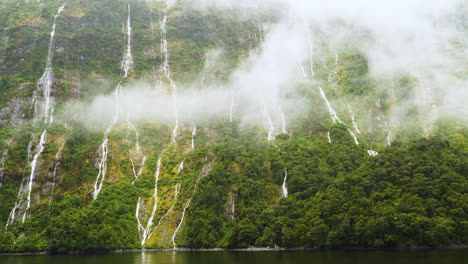 Cascade-waterfalls-flowing-from-cliff-in-milford-sound,-over-mist-evaporating-from-rain-forest