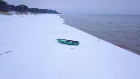 Aerial-view-of-Baltic-sea-coast-on-a-overcast-winter-day-with-green-coastal-fisherman-boat,-beach-with-white-sand-covered-by-snow,-coastal-erosion,-wide-angle-orbiting-drone-shot