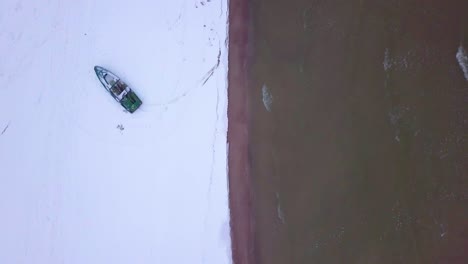 Aerial-birdseye-view-of-Baltic-sea-coast-on-a-overcast-winter-day-with-green-coastal-fisherman-boat,-beach-with-white-sand-covered-by-snow,-coastal-erosion,-wide-angle-drone-shot-moving-forward