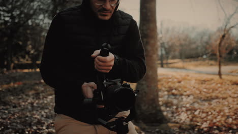 Happy-and-cheerful-young-caucasian-male-videographer-wearing-glasses,-a-black-vest,-and-a-hoodie,-using-a-video-camera-and-stabilizer-to-record-the-autumn-season-in-a-forest-out-in-nature