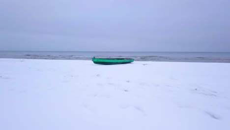 Aerial-view-of-Baltic-sea-coast-on-a-overcast-winter-day-with-green-coastal-fisherman-boat,-beach-with-white-sand-covered-by-snow,-coastal-erosion,-wide-angle-drone-shot-moving-forward-low