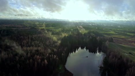 Forestry-landscape-with-lake-water-and-low-flowing-clouds,-aerial-view