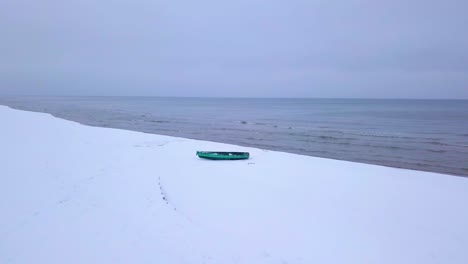 Aerial-view-of-Baltic-sea-coast-on-a-overcast-winter-day-with-green-coastal-fisherman-boat,-beach-with-white-sand-covered-by-snow,-coastal-erosion,-wide-angle-drone-shot-moving-forward-low
