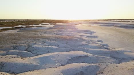 moving-aerial-view-of-a-salt-field,-snowy-field-with-little-vegetation-and-a-sunset-sky