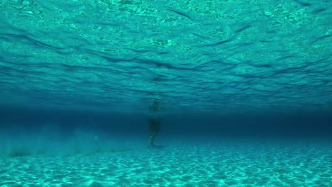 Wunderschöne-Unterwasserszene-Von-Menschen,-Die-Auf-Sandigem-Meeresboden-Spazieren-Gehen,-Mit-Sonnenlichtbrechungen-Und-Reflexionen-In-Türkisfarbenem,-Transparentem-Meerwasser
