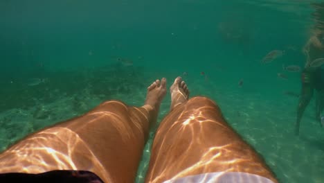Underwater-personal-perspective-view-of-man-legs-floating-in-clear-transparent-sea-water-with-people-bathing-in-background,-slow-motion