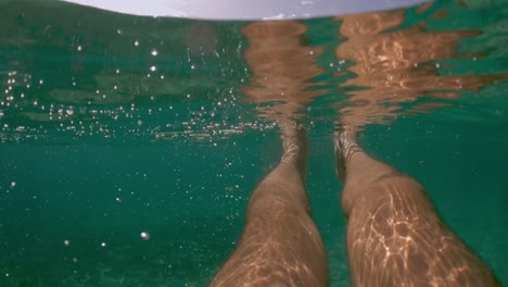 Underwater-personal-perspective-view-of-man-legs-floating-in-clear-transparent-sea-water-with-waterline-edge-and-coast-in-background,-slow-motion