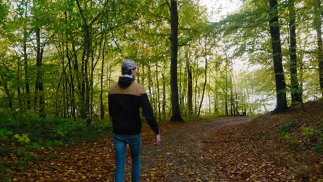 Young-Man-WIth-Cap-and-Jacket-Walks-on-a-Small-Path-in-Gyllebo-Forest-in-Auyumn-Afternoon,-Österlen-Sweden---Tracking-Medium-Wide-Shot-From-behind