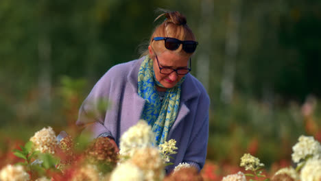 Eine-Frau-Im-Mantel,-Die-Eine-Tasche-Mit-Brille-Trägt,-Geht-Durch-Einen-Blumengarten,-Während-Sie-Im-Sommer-In-Litauen-Die-Blumenknospen-Riecht
