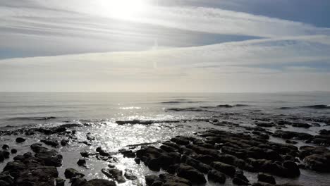 Aerial-Low-Flying-Over-Silhouette-Of-Rocks-With-Waves-Gently-Crashing-Over-On-Sunny-Bright-Day
