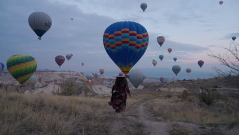 Girl-Running-Towards-Scenic-Pathway-In-A-Field-In-Cappadocia-To-Witness-Marvelous-Hot-Air-Balloons-In-Flight---Wide-shot