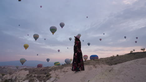 Frau-In-Langem-Kleid,-Die-Auf-Einem-Berg-Steht-Und-Bunte-Hor-luftballons-Im-Flug-In-Kappadokien,-Türkei-Bei-Sonnenaufgang-Betrachtet