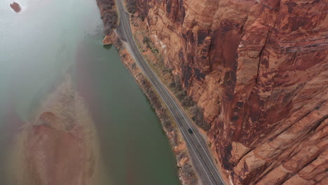 Aerial-View-of-Black-Vehicle-on-Deserted-Road-by-Colorado-River-Under-High-Steep-Cliff-in-Utah-Landscape,-Tilt-Down-Drone-Shot
