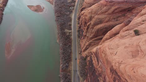 Birds-Eye-Aerial-View-of-Lonely-Car-on-Deserted-Road-by-Colorado-River-Under-Steep-High-Sandstone-Cliffs-of-Utah-Desert,-Tracking-Drone-Shot