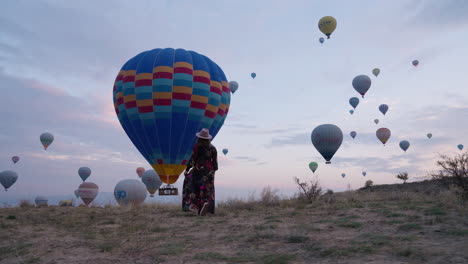 Girl-In-Dress-Walking-In-A-Field-Overlooking-Beautiful-Hot-Air-Balloons-In-Flight-In-Cappadocia---panning-shot