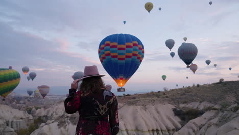 Female-Tourist-In-Long-Dress-And-Hat-Watching-Colorful-Hot-Air-Balloons-Flying-In-The-Sky-At-Sunrise-In-Cappadocia,-Turkey