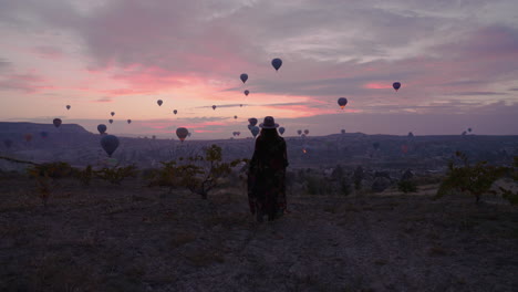 Lady-In-Dress-Walking-Outdoor-To-View-Stunning-Hot-Air-Balloons-Scenery-In-The-Sky-At-Dawn-In-Cappadocia,-Turkey,-wide,-following-shot