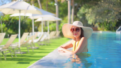 A-pretty-woman-standing-in-a-swimming-pool-wearing-a-huge-white-sun-hat-and-sunglasses-smiles-as-she-makes-eye-contact-with-the-camera