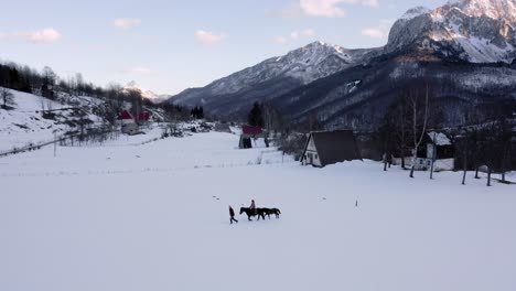 AERIAL---People-riding-a-horse-on-a-farm-on-a-snowy-winter,-Kolasin,-Montenegro,-pan-left