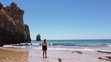 Playa-Praia-Dos-Tres-Irmaos,-Algarve,-Portugal---Chica-Turista-Caminando-En-La-Playa-De-Arena-A-Través-Del-Mar-Azul-Claro